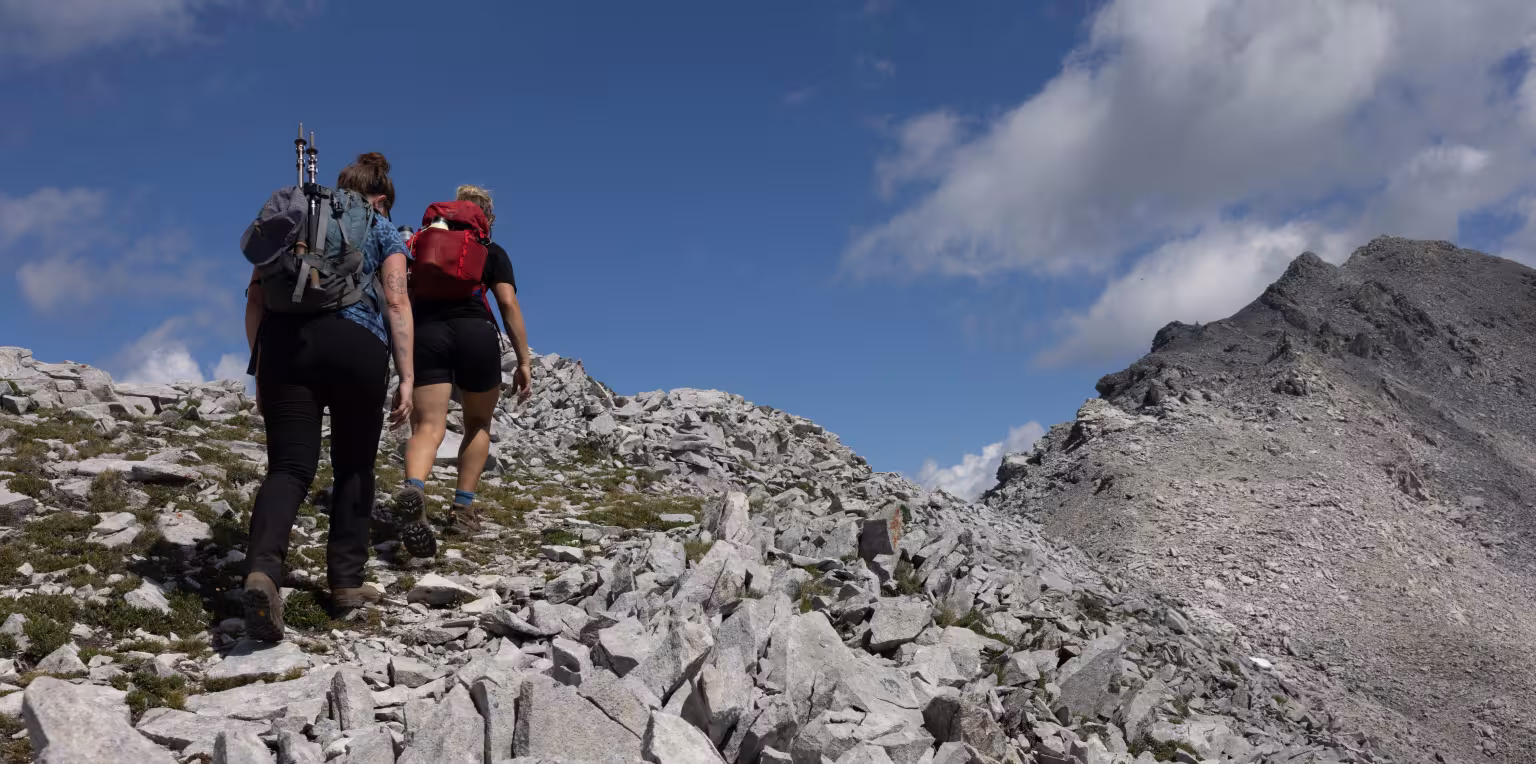 Two hikers on a rocky mountain trail, blue sky with clouds behind.