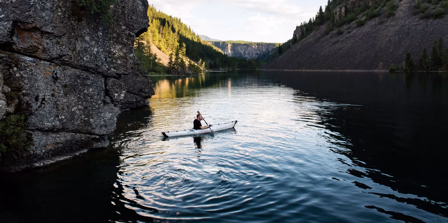 Person canoeing on calm river surrounded by steep cliffs and trees.