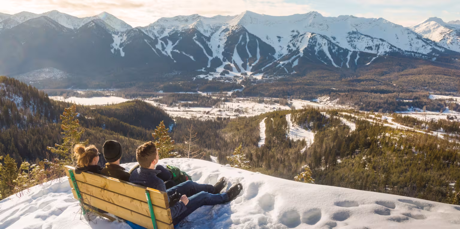 Three people sitting on wooden bench at the Hyperventilation viewpoint overlooking snowy mountains.