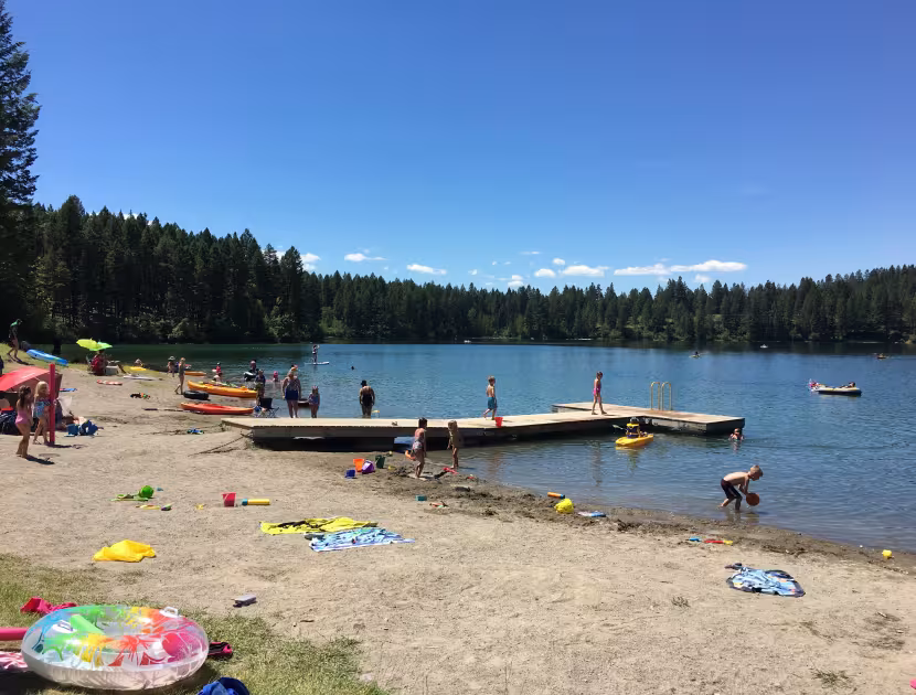 Beach scene with people, kayaks, and inflatables on shore, dock in lake, trees in background.