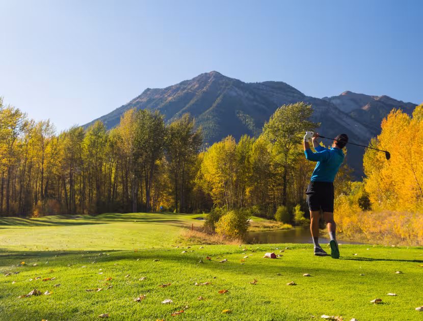 Golfer in blue shirt swings club on green course with mountains behind.