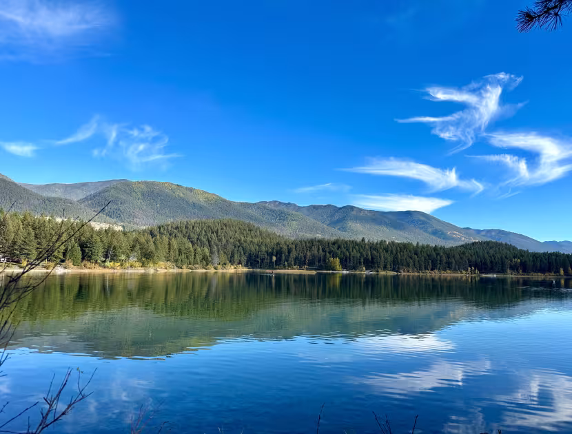 Serene lake reflecting mountains and trees under a blue sky with wispy clouds.