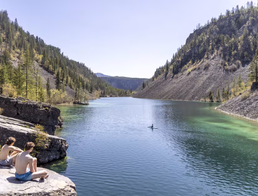 Two men sitting on rocky cliffside overlooking serene lake surrounded by mountains and trees.