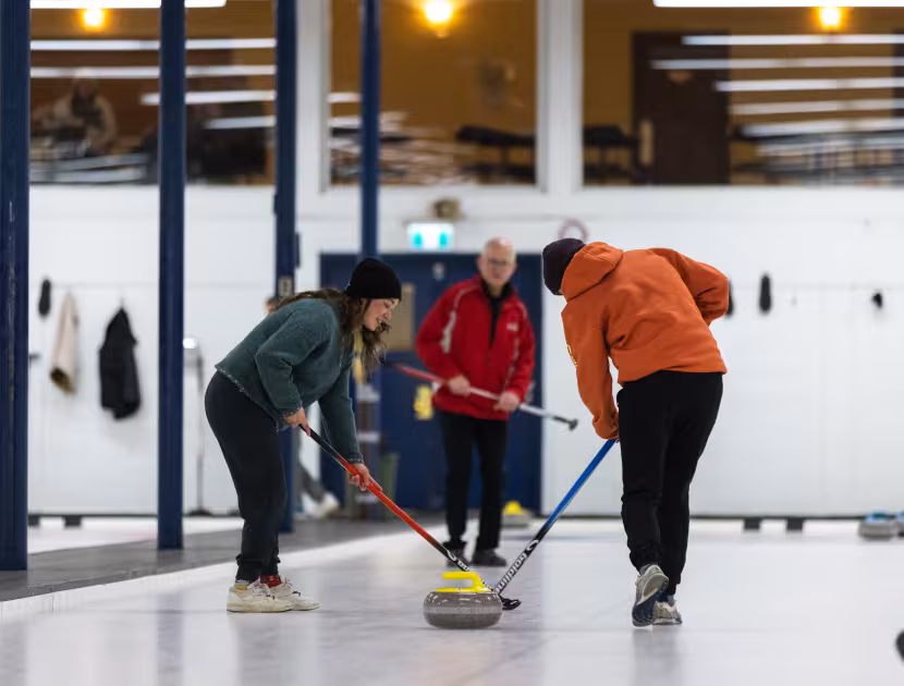 Two people sweeping ice with brooms, one wearing orange, the other green; third person in background.