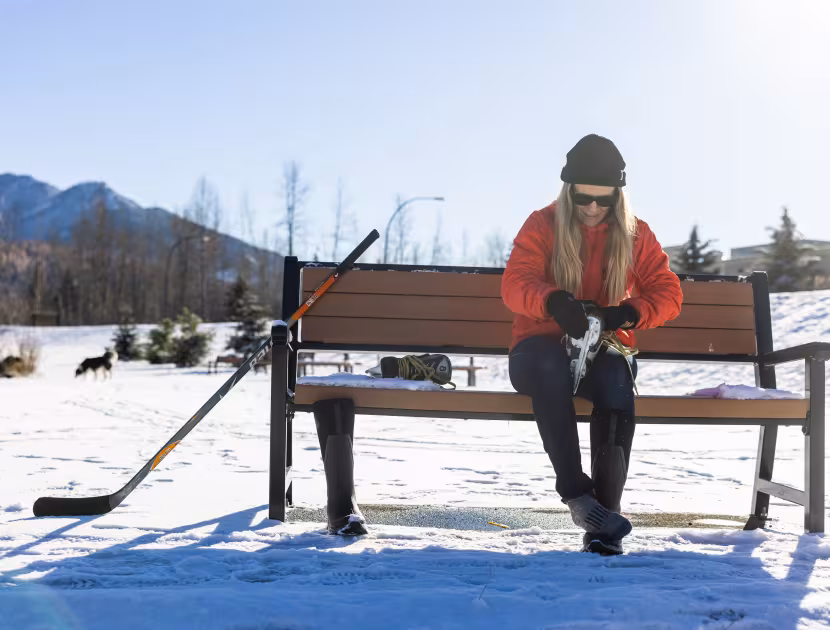 Woman in orange jacket sitting on a bench with a hockey stick beside her in a snowy landscape.