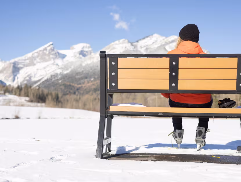 Person in orange jacket sitting on a bench, looking at snowy mountains.