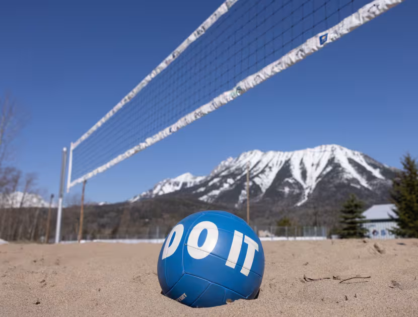 Blue volleyball with white text on sandy court, snow-capped mountains in background.