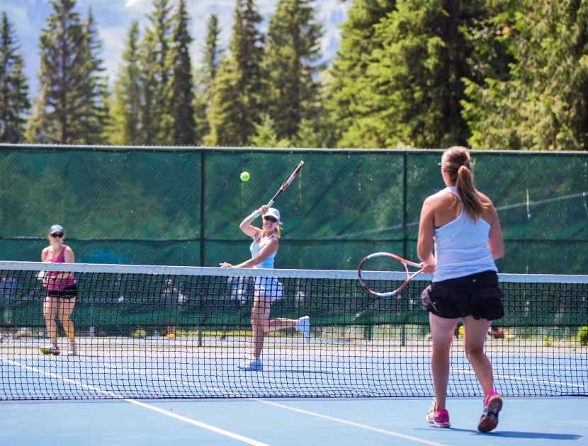 Three women on outdoor tennis court, one hitting ball with racket; green trees and fence behind.