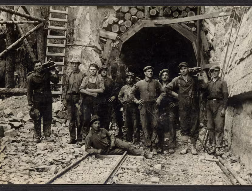 Group of men in old-fashioned clothing stand in front of a mine entrance, one sitting in front with a log.