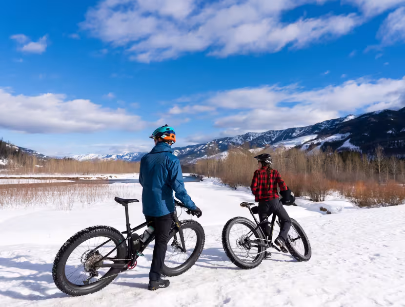 Two people in winter gear standing with fat bicycles on snowy trail, mountains in background.