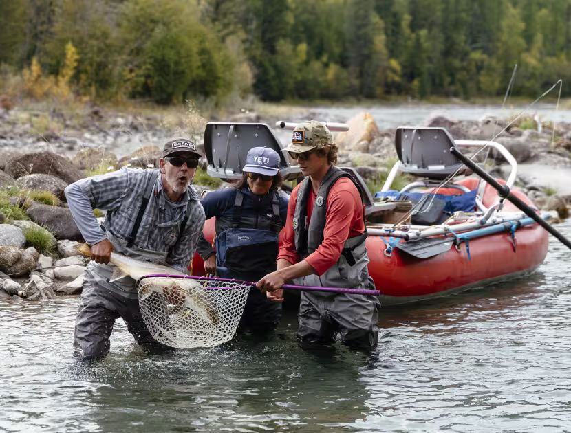 Three men in waders holding a net and fishing gear in a river, with a red raft behind them.