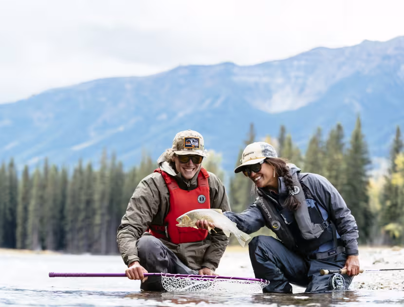 Two people crouching in shallow water, holding a large fish and a fishing net, with mountains and trees in background.