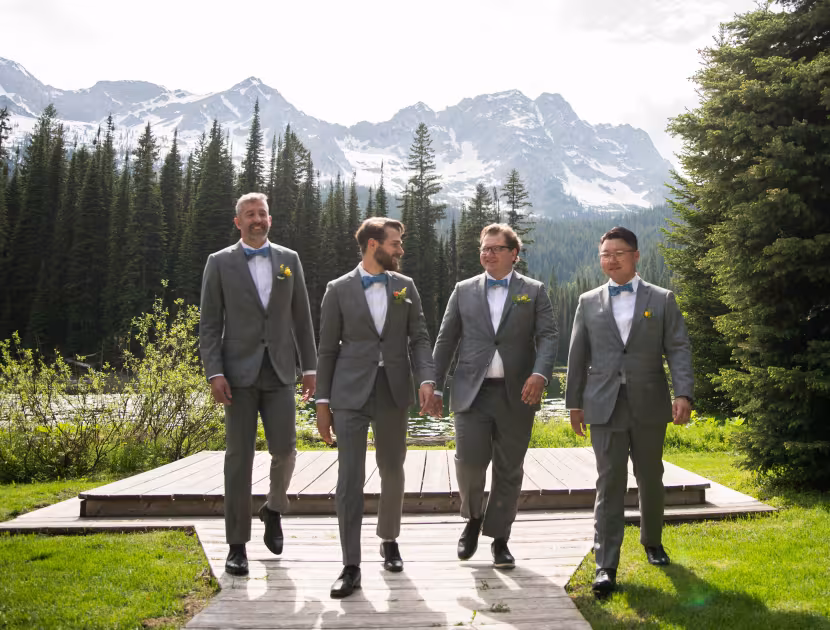 Four men in grey suits standing on a wooden platform outdoors, mountains in background.