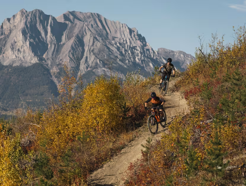 Two mountain bikers riding on a dirt trail with a large mountain in the background.