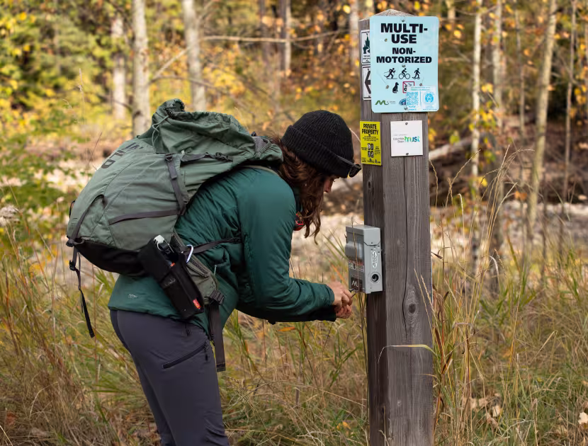 Woman in green jacket and backpack interacting with trail register box on wooden post.