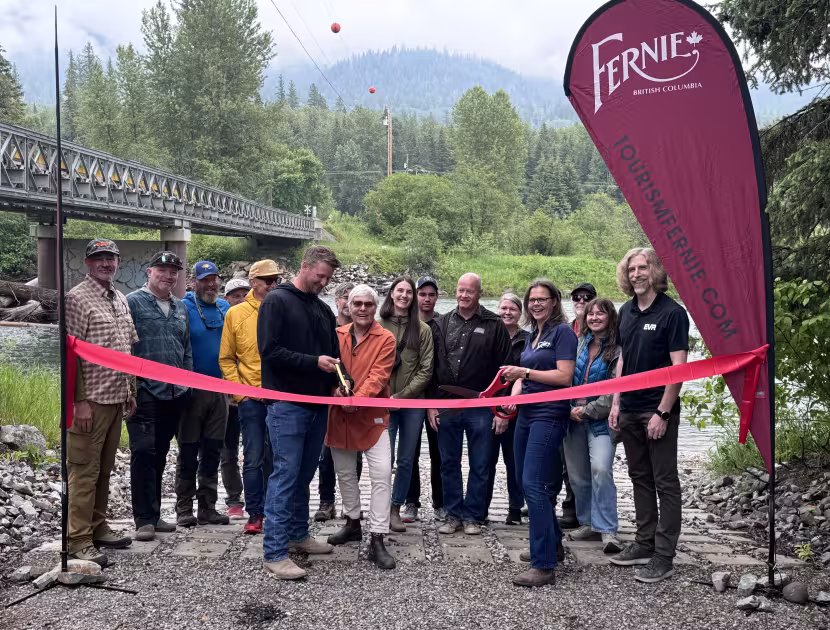 Group of people at a ribbon cutting ceremony outdoors, holding a long pink ribbon; a red flag with 'Fernie' on it stands to the right.