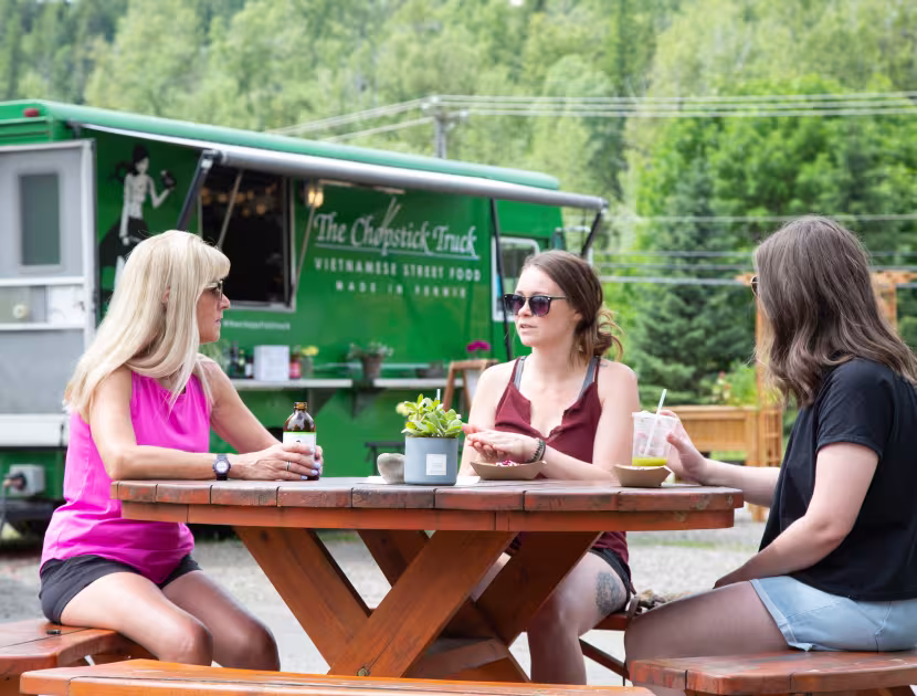 Three women sitting at a picnic table outside a green food truck.