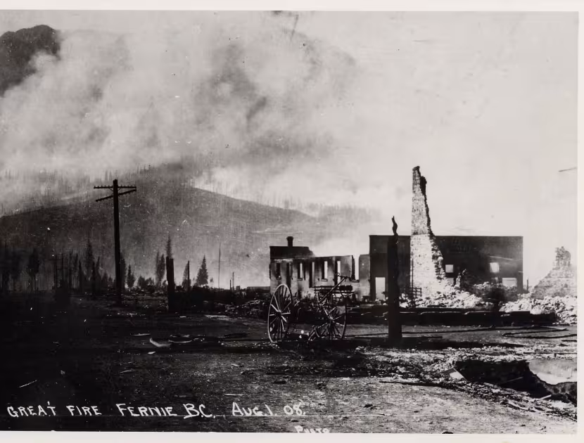 Black and white photograph, a smoky industrial area with buildings and a crane; dark hills in the background.