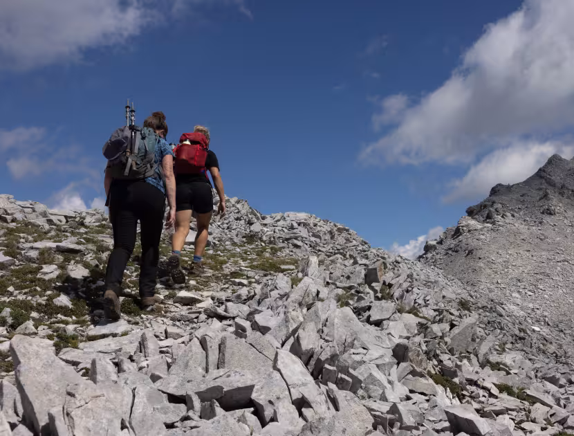 Two hikers on a rocky mountain trail, blue sky with clouds behind.