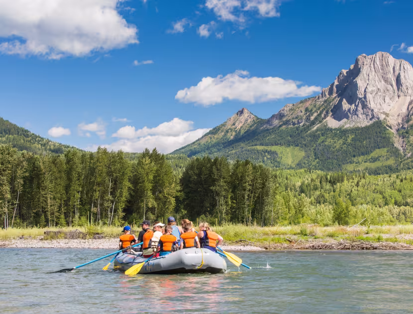 People in orange life jackets on a raft in a river, with mountains and trees in the background.