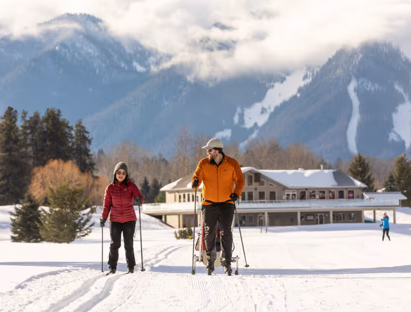 Two cross-country skiers, one towing a baby chariot on a snowy field with a large building and mountain range in the background.