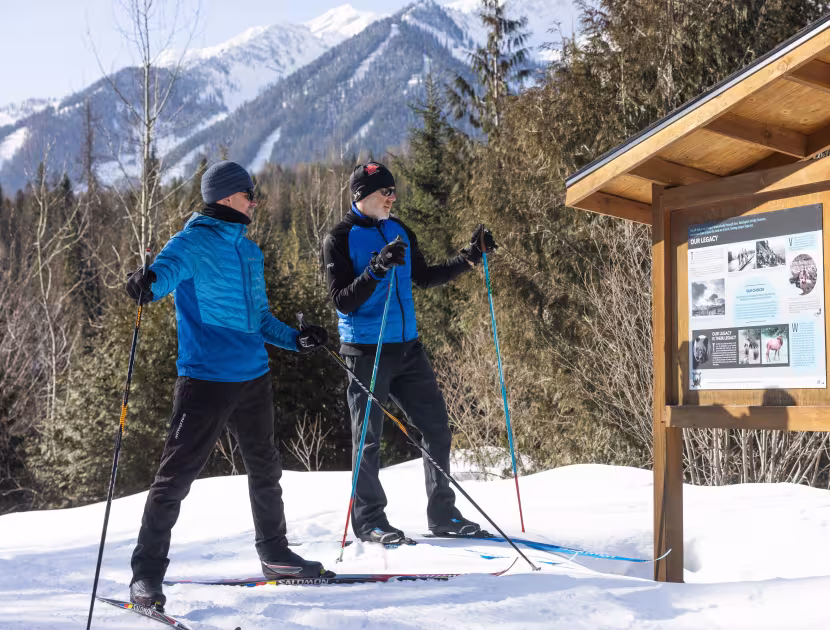 Two cross-country skiers in blue jackets and black pants on snow, wooden sign with information board beside them, snowy mountains and trees background.
