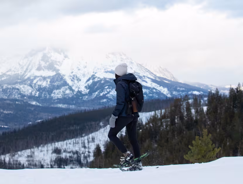 Person snowshoeing on a snowy slope with a mountain range and trees in the background.