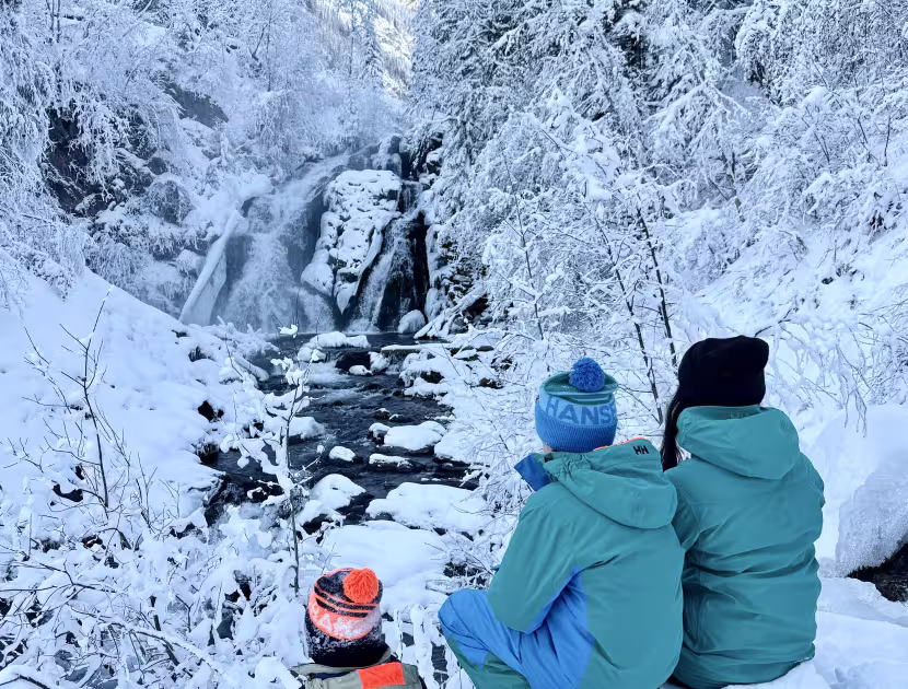 Two people in teal coats sitting on snowy bank, looking at frozen waterfall; person on left wears blue hat, person on right wears black hat.