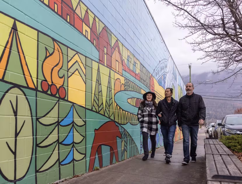 Three people walking on sidewalk past colorful outdoor mural depicting camping scenes.