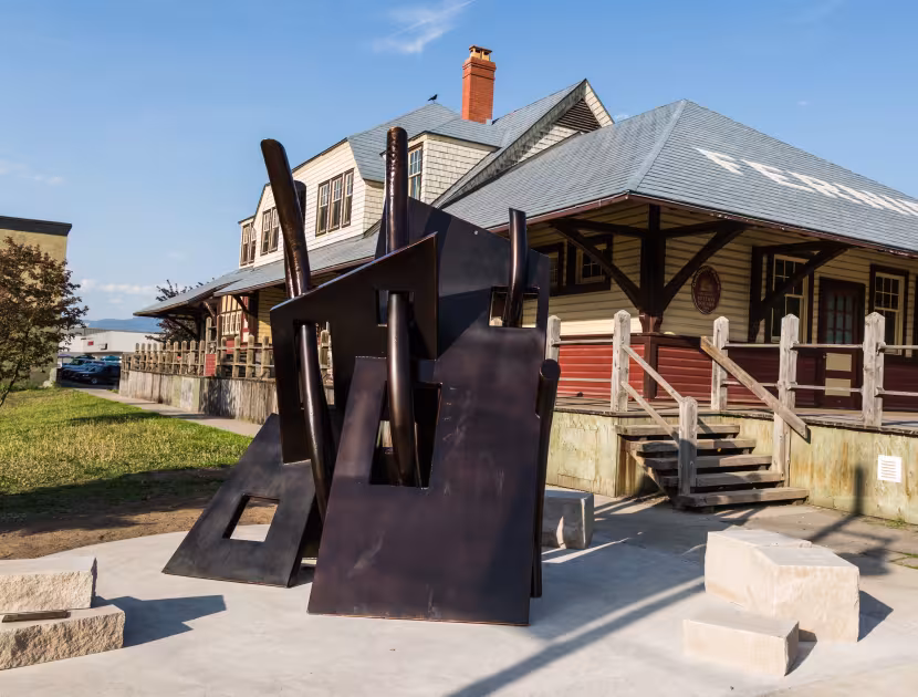 A large abstract metal sculpture in front of a small house with a porch and stairs, under a blue sky.