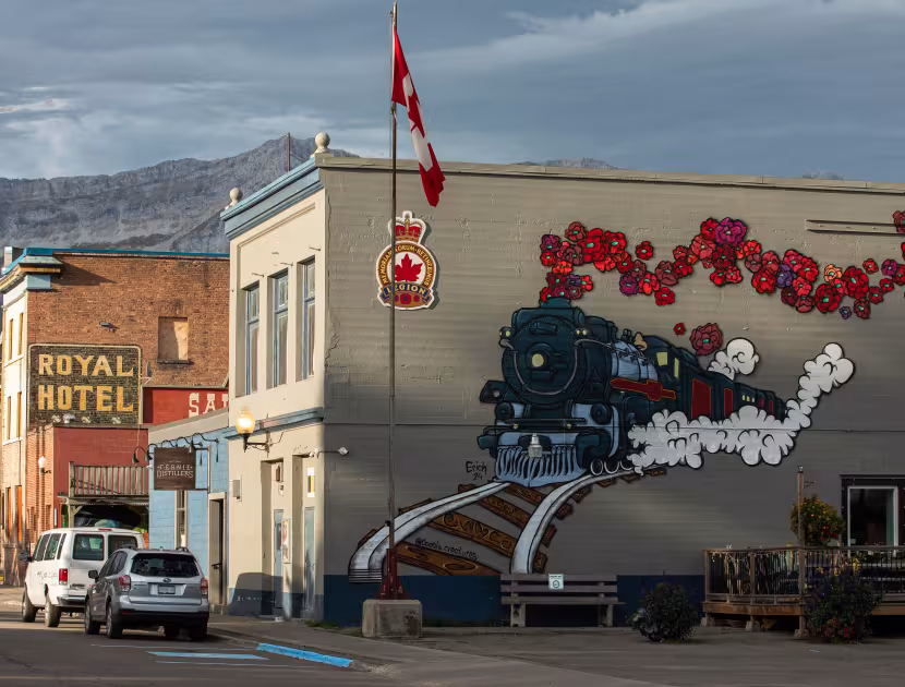 A mural on a beige building depicting a train with a red and white trail; mountains in background; Canadian flag flying.