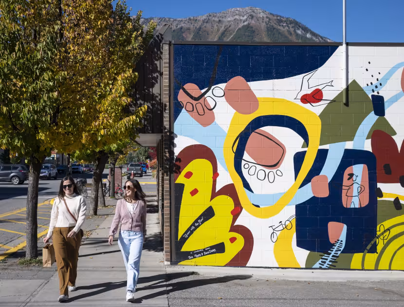 Two women walking on a sidewalk past a colorful mural with a mountain in the background.
