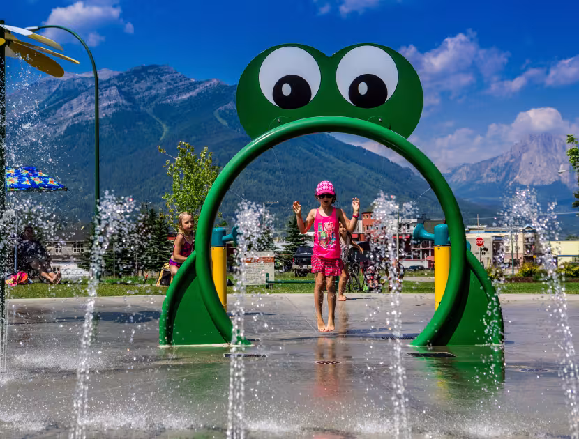 Child in pink plays under frog-shaped water feature at splash pad with mountains in background.