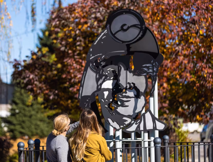 Two people looking up at a large mural of a man's face; trees with fall foliage in background.