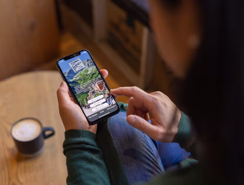 Woman sitting at a table, holding smartphone with the Iconic Fernie mobile app on the screen.