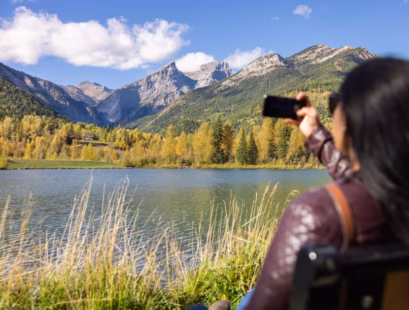 Woman sitting on a bench taking photo of lake and mountains with phone.