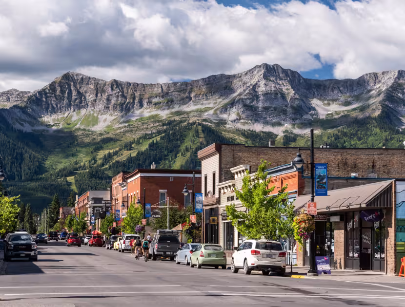A small town's main street lined with shops and cars, surrounded by green mountains under a cloudy sky.