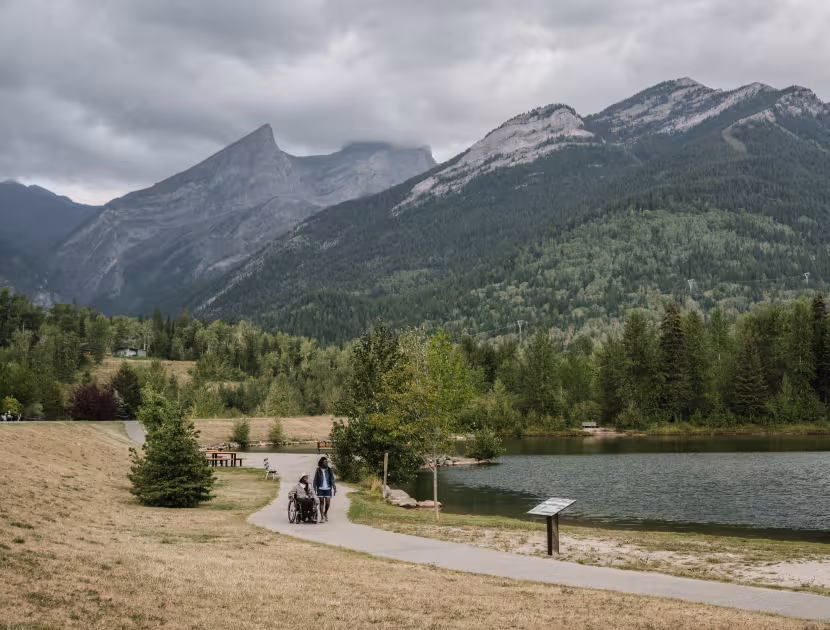Serene lake surrounded by lush greenery and mountains under a cloudy sky, with a walking path and 2 people in the distance, one of them in a wheelchair..