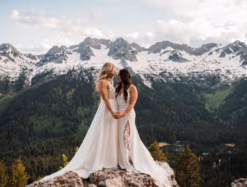 Two brides in white wedding dresses embracing on a rocky outcrop, overlooking a mountainous landscape.