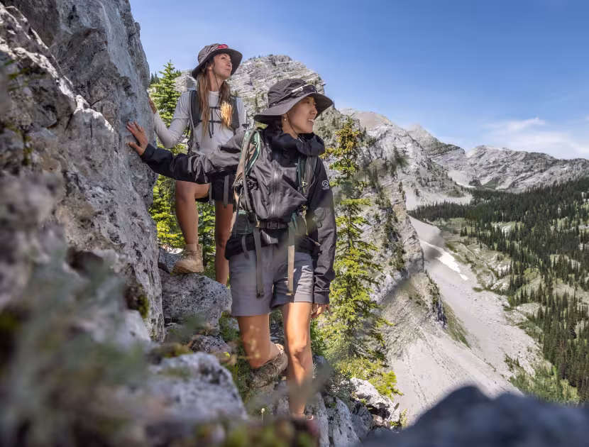Two hikers on a rocky mountain trail, looking out at a scenic valley with trees and mountains.
