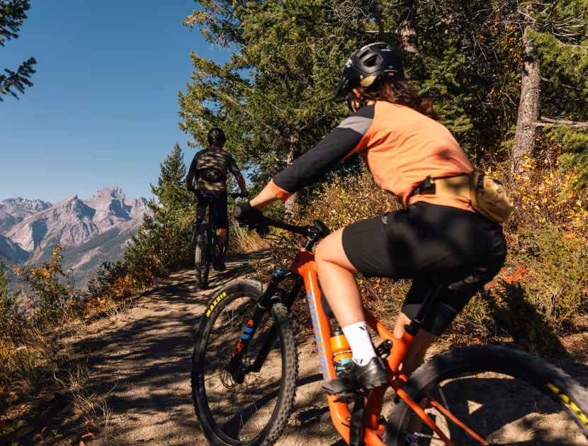 Two mountain bikers riding on a trail in fall with mountains in the background.