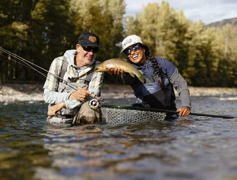 Two anglers in a river, holding a large fish and fishing rods, smiling; trees and mountains in background.