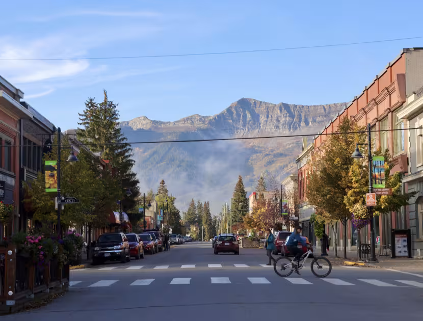 Mountainous town street with cyclist, trees, and buildings; blue sky.