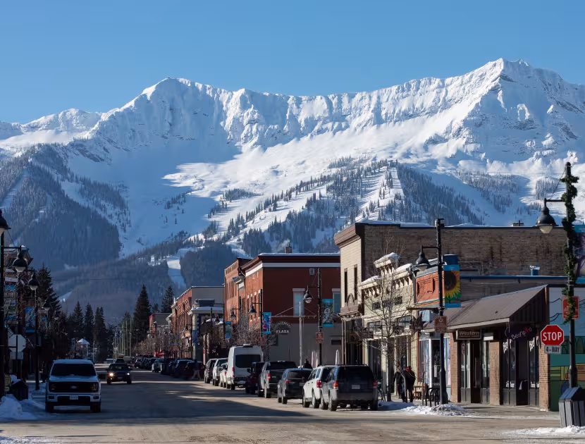 Snowy mountain range behind a small town's main street with parked cars and buildings.