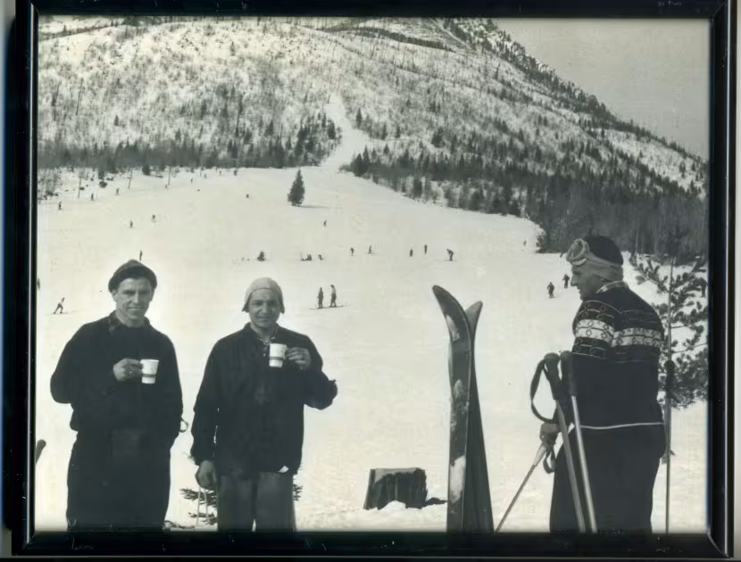A historic black & white photograph of three people in winter clothing standing on a snowy slope, with a snowy hill and trees in the background.