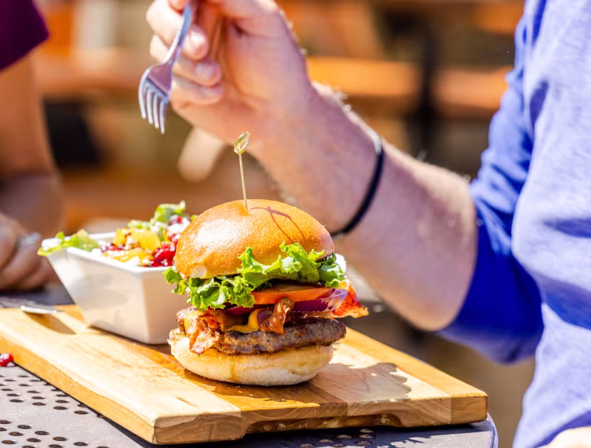 Person in blue shirt holding a fork over a burger on a wooden board with a side dish.