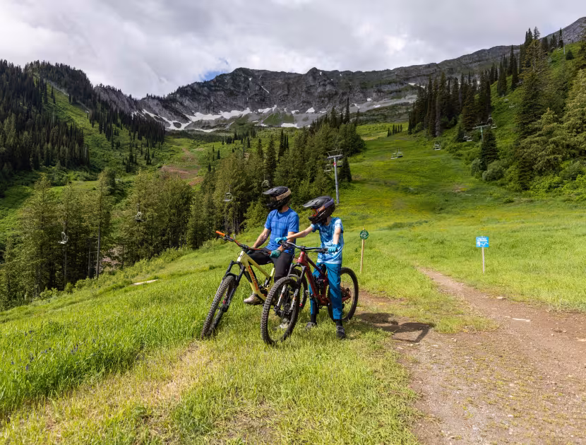 Two mountain bikers standing with bikes on a grassy trail, mountains and trees in background.