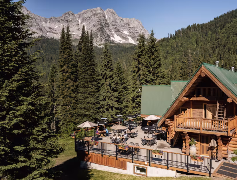 Log cabin with green roof and a patio with tables and umbrellas surrounded by evergreen trees and mountain range.