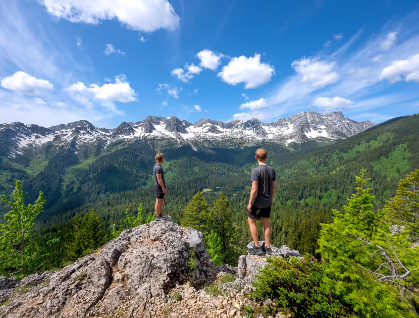 Two people stand on a rocky outcropping overlooking a mountainous landscape with green trees and snow-capped mountains under a blue sky with clouds.