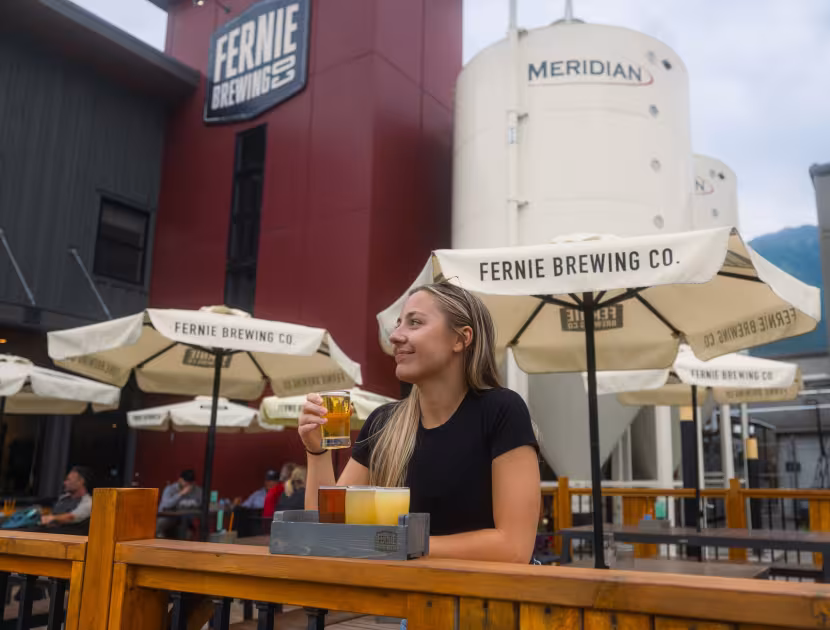 Woman sitting at outdoor patio table, holding a beer.